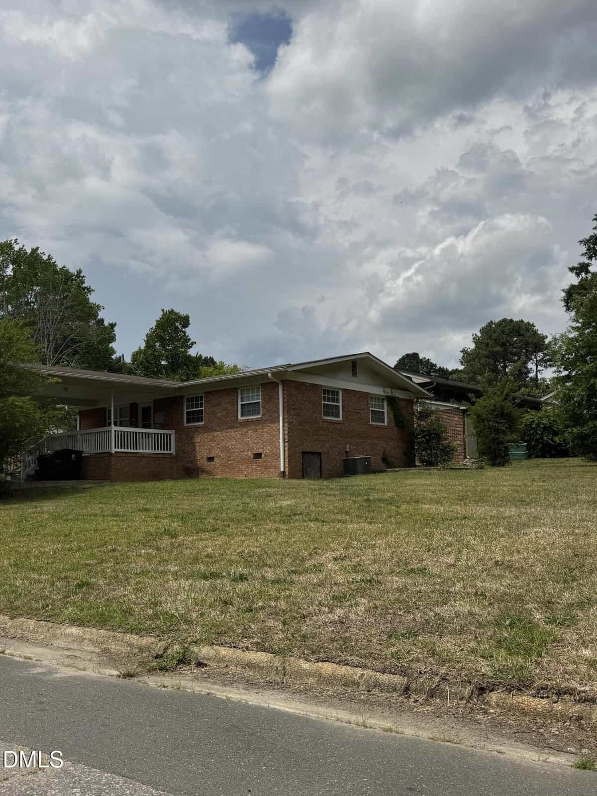 A single-story brick house with a covered carport is set on a slightly sloped yard with patchy grass. Trees and shrubbery border the property, and the sky is partly cloudy. A DMMLS watermark is in the bottom left corner.