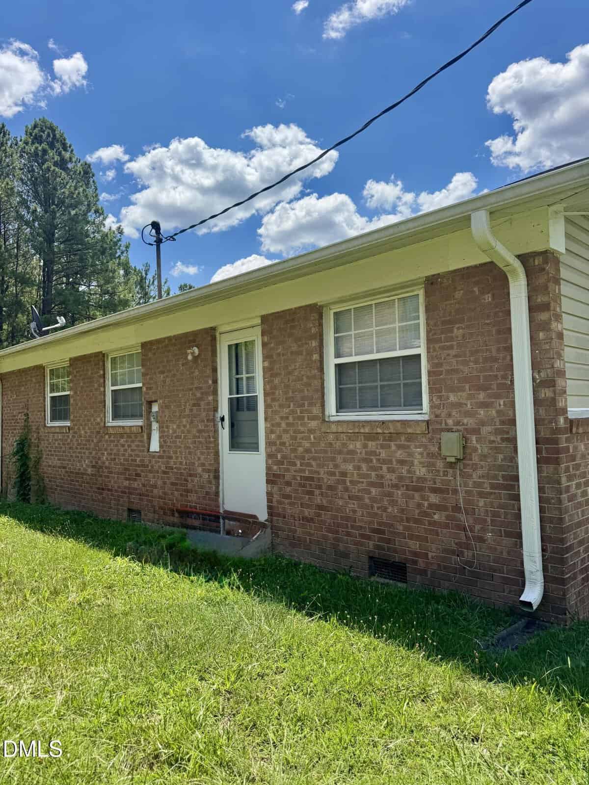 A single-story brick house with a white back door, two windows, a rain gutter, and a grassy yard. The sky is blue with scattered clouds, and pine trees are visible in the background.