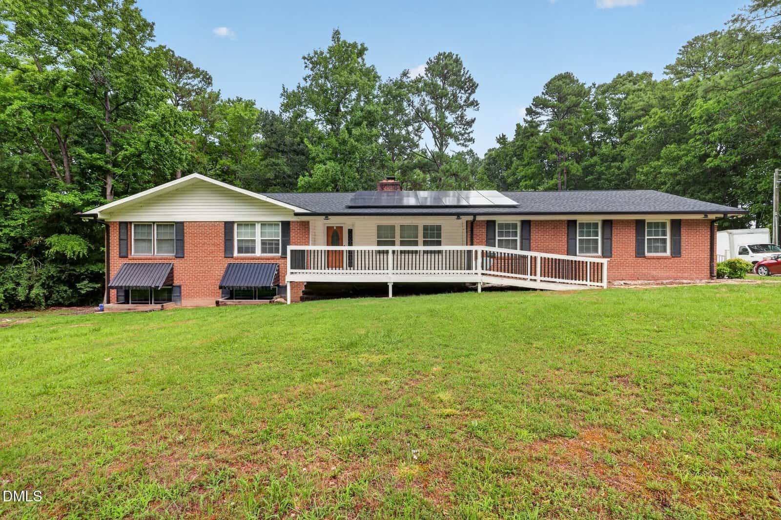 Charming single-story red brick house at 56 Lee Lane, Henderson, featuring a black roof with solar panels, crisp white trim, and a white accessibility ramp leading to the front door. The lawn is green with some patchy areas, while mature trees provide a picturesque backdrop.