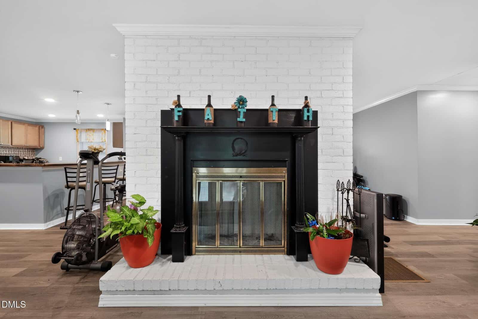 A white brick fireplace with a black mantle, decorated with “FAITH” letters on top. Two red plant pots sit on the hearth, and a dining area and kitchen are visible in the background.