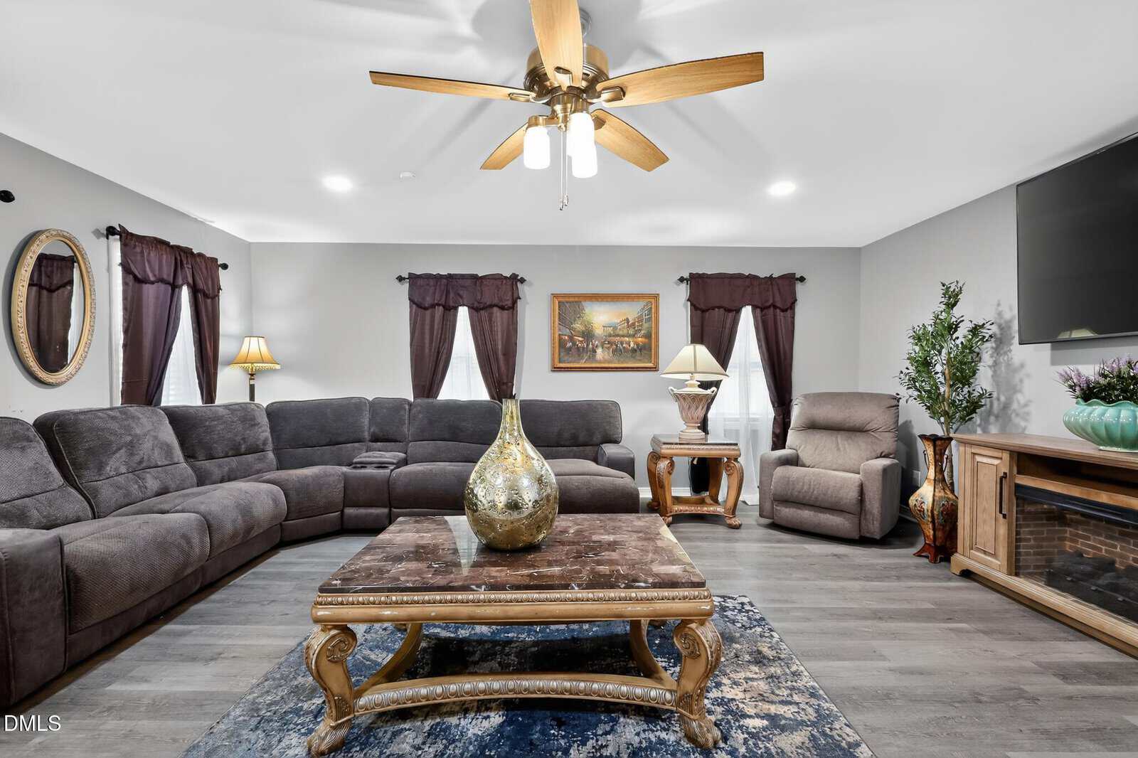 A living room with gray sectional sofa, recliner, wooden coffee table with large gold vase, dark curtains, lamps, framed painting, TV, fireplace, and potted plant on hardwood floors with a blue patterned rug.