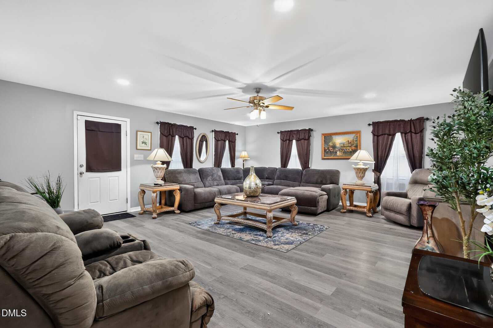 A spacious living room with gray sofas, a patterned area rug, wood tables with lamps, a ceiling fan with lights, wall art, and windows with dark curtains. A TV is on a stand to the right. The floor is gray wood.