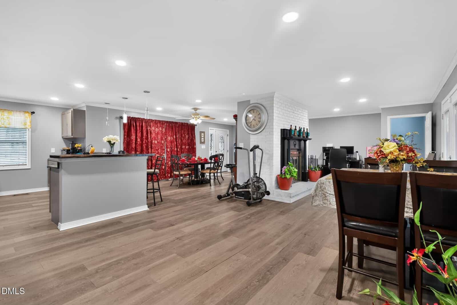 Open-plan living space with a kitchen, dining area, and living room. Features gray walls, wood flooring, an exercise bike, red curtains, white brick fireplace, and wooden furniture, with plants and home decor throughout.