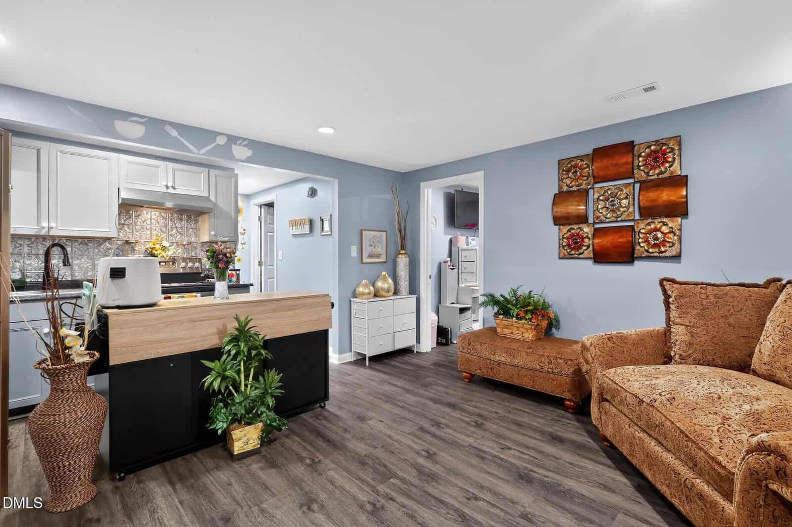 A cozy living room with a brown sofa, wooden flooring, blue walls, plants, wall art, and an open view into a white kitchen with modern decor.