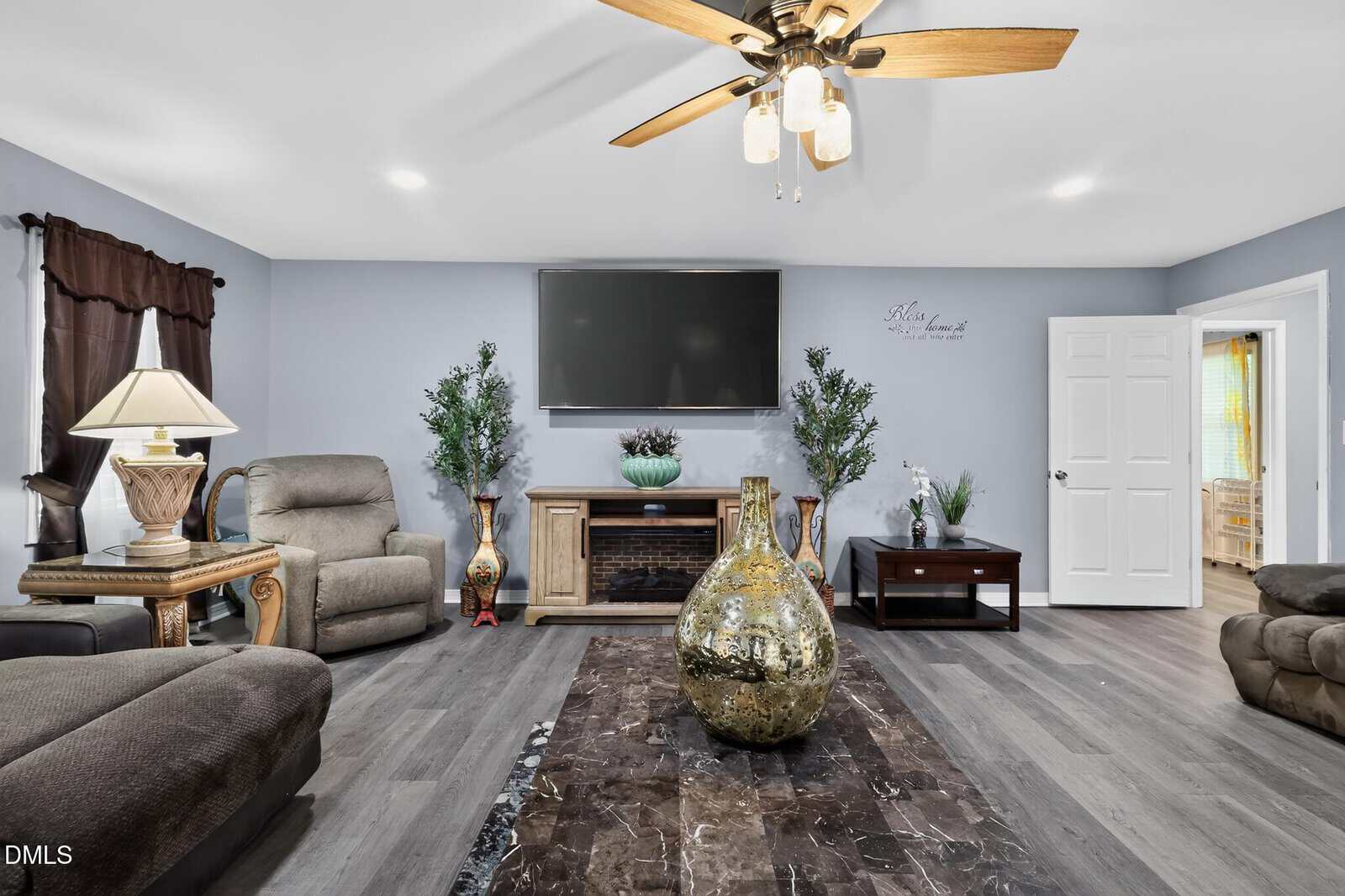 Modern living room with gray walls, wood floors, a ceiling fan, large wall-mounted TV, marble coffee table, brown sofas, recliner, two potted plants, and decorative items. Door open to another room on the right.
