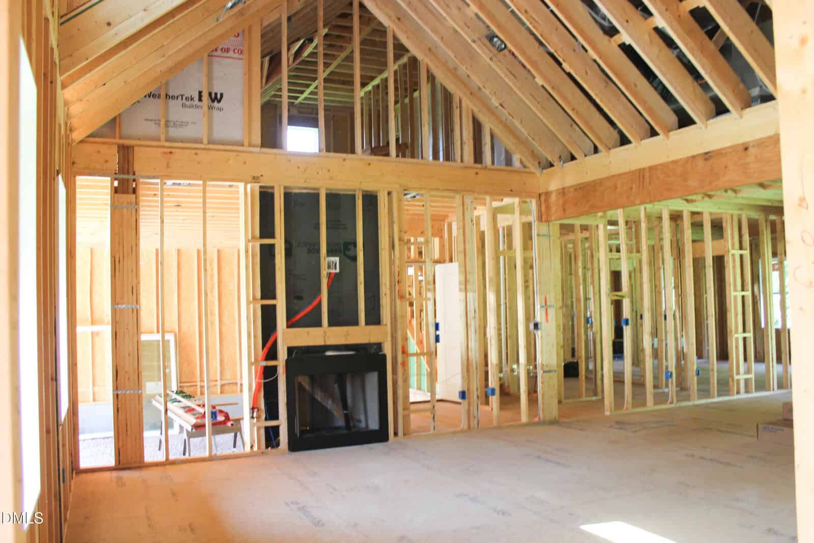 Interior view of a house under construction, showing exposed wooden framing, insulation, and an unfinished fireplace. No drywall or finishing materials are present. Natural light enters through window openings.