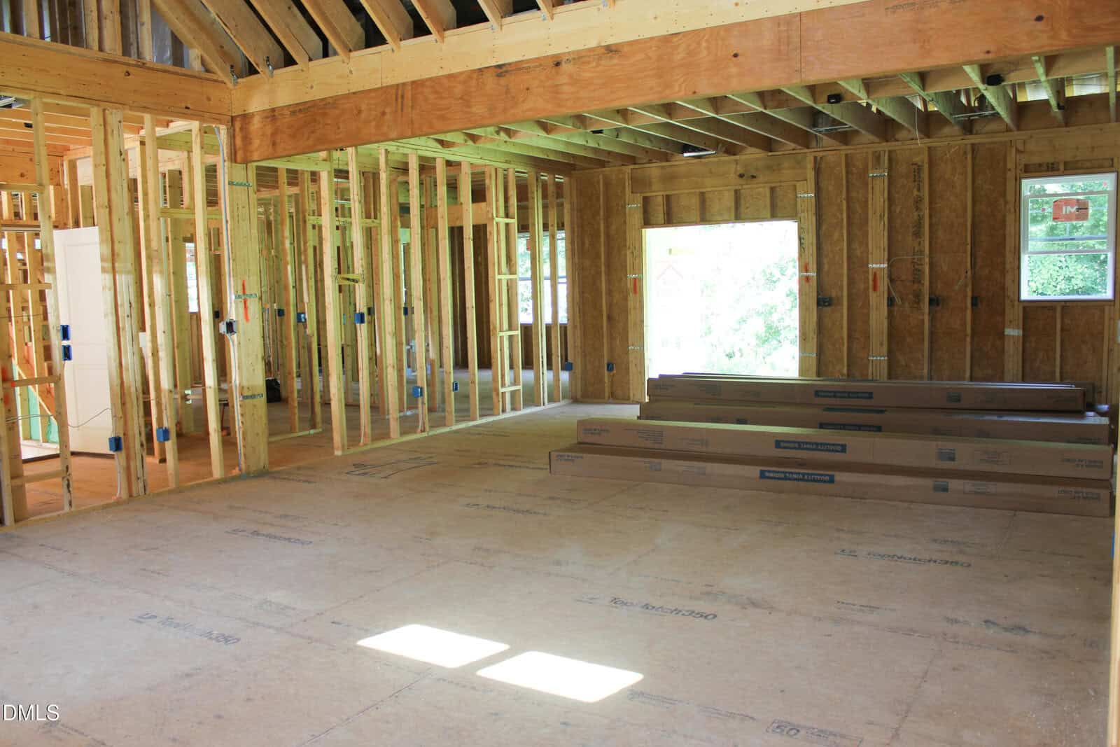 A house under construction with exposed wooden framing, an unfinished floor, and stacks of building materials. Sunlight comes through an open window and a large wall opening. No finished walls or fixtures are present.
