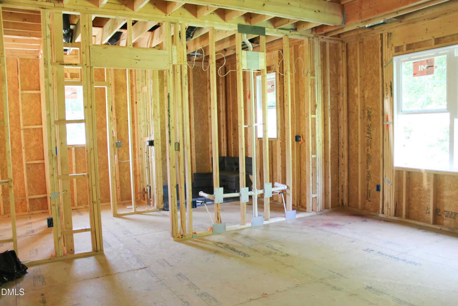 Unfinished room under construction with exposed wooden framing, plumbing, and electrical wiring. Sunlight comes in through a window on the right, illuminating the partially built interior.