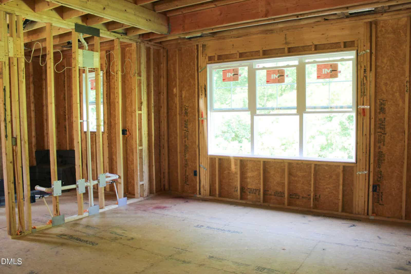 Unfinished room under construction with exposed wooden framing, electrical wiring, and a large window letting in natural light. No drywall or flooring installed yet.