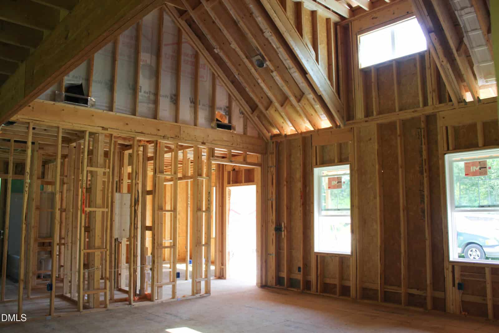 Interior view of a house under construction showing exposed wooden framing, insulation in the ceiling, unpainted walls, multiple windows, and natural light entering the room. No finished fixtures or flooring are present.