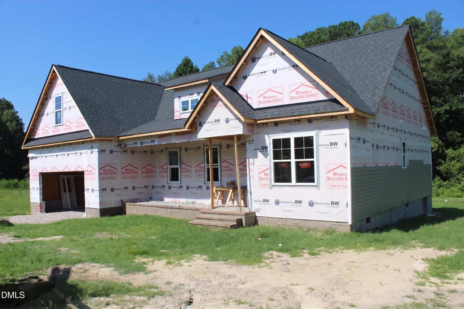 A partially constructed house with visible insulation wrap, unfinished porch steps, and some siding installed. The roof is complete, and the house is surrounded by grass and trees under a clear sky.
