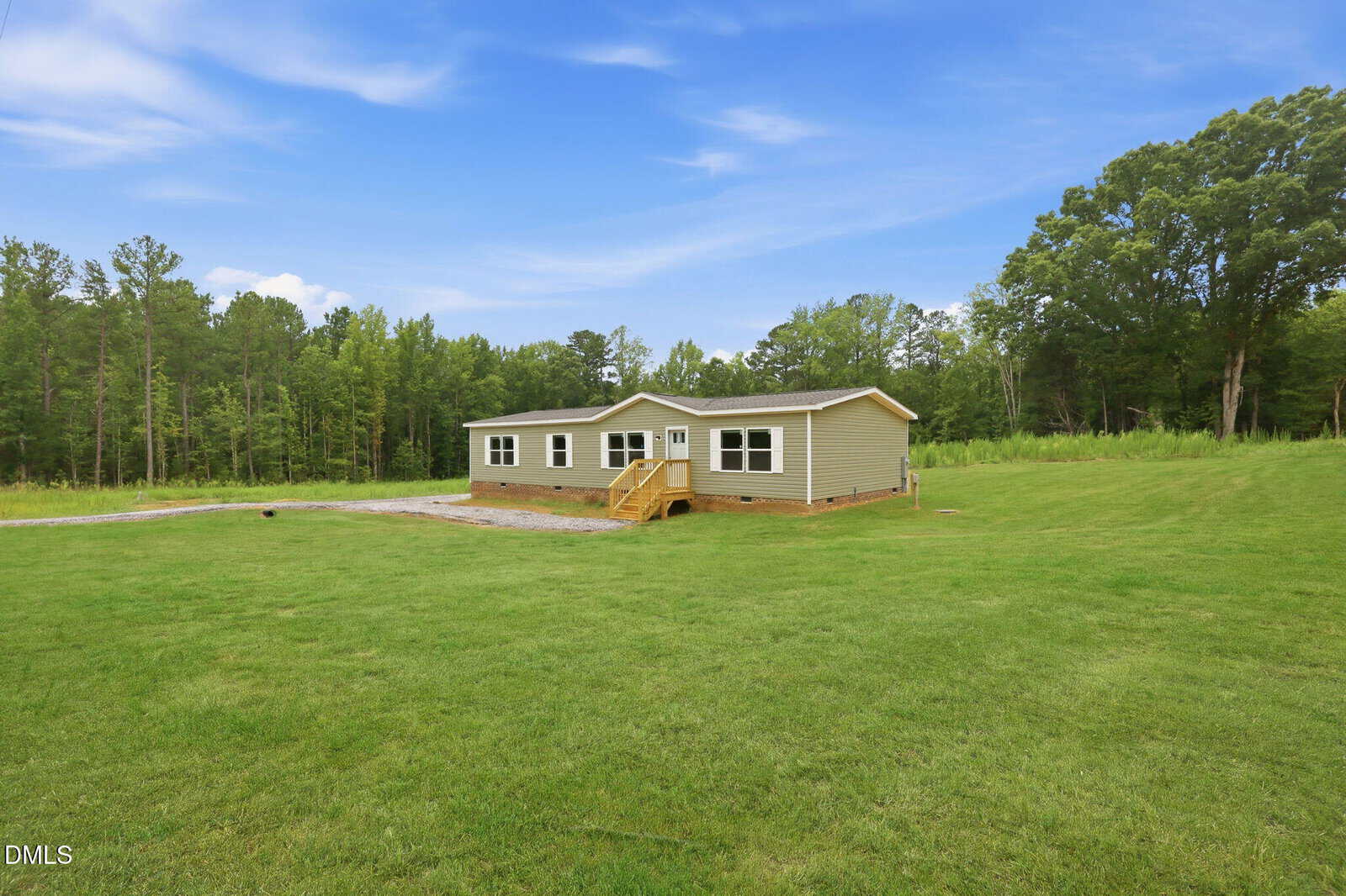 A single-story, beige house with a small porch and steps sits on a spacious grassy lawn, surrounded by trees under a partly cloudy blue sky.