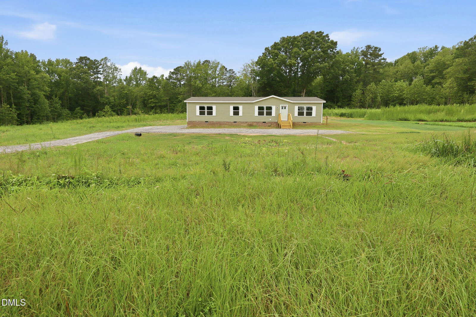 A single-story manufactured home with light-colored siding sits on a large grassy lot, surrounded by trees and a gravel driveway, under a partly cloudy sky.