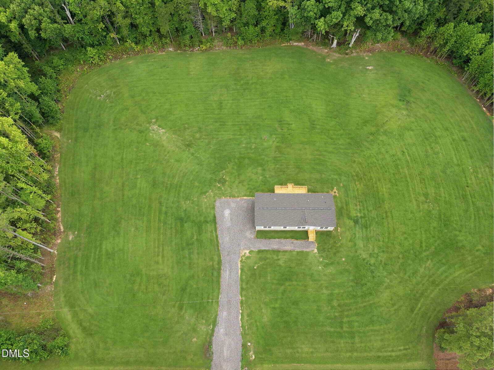Aerial view of a single-story house with a gray roof, surrounded by a large, neatly mowed grassy lawn and bordered by dense green trees. A gravel driveway leads to the house and there is a small wooden deck at the entrance.