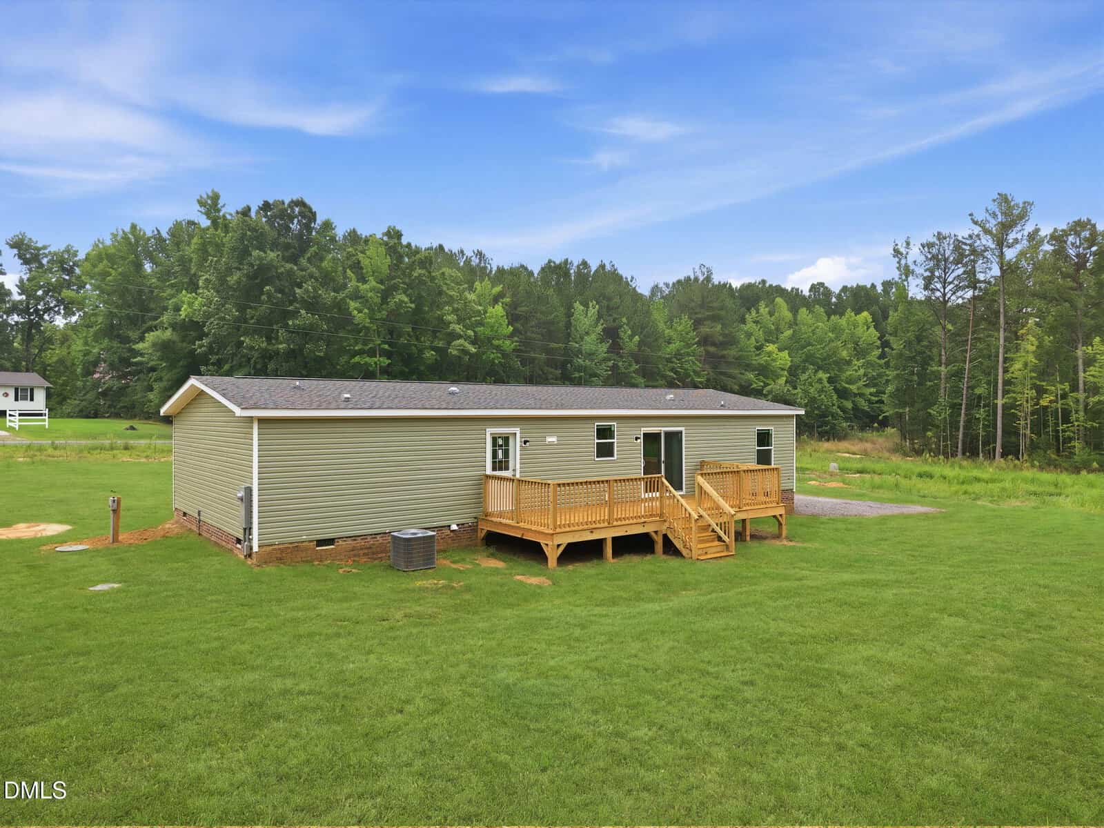 A single-story beige house with a gray roof and wooden deck is centered in a grassy yard. The background features green trees and a partly cloudy sky. The house has multiple windows and a simple, modern design.