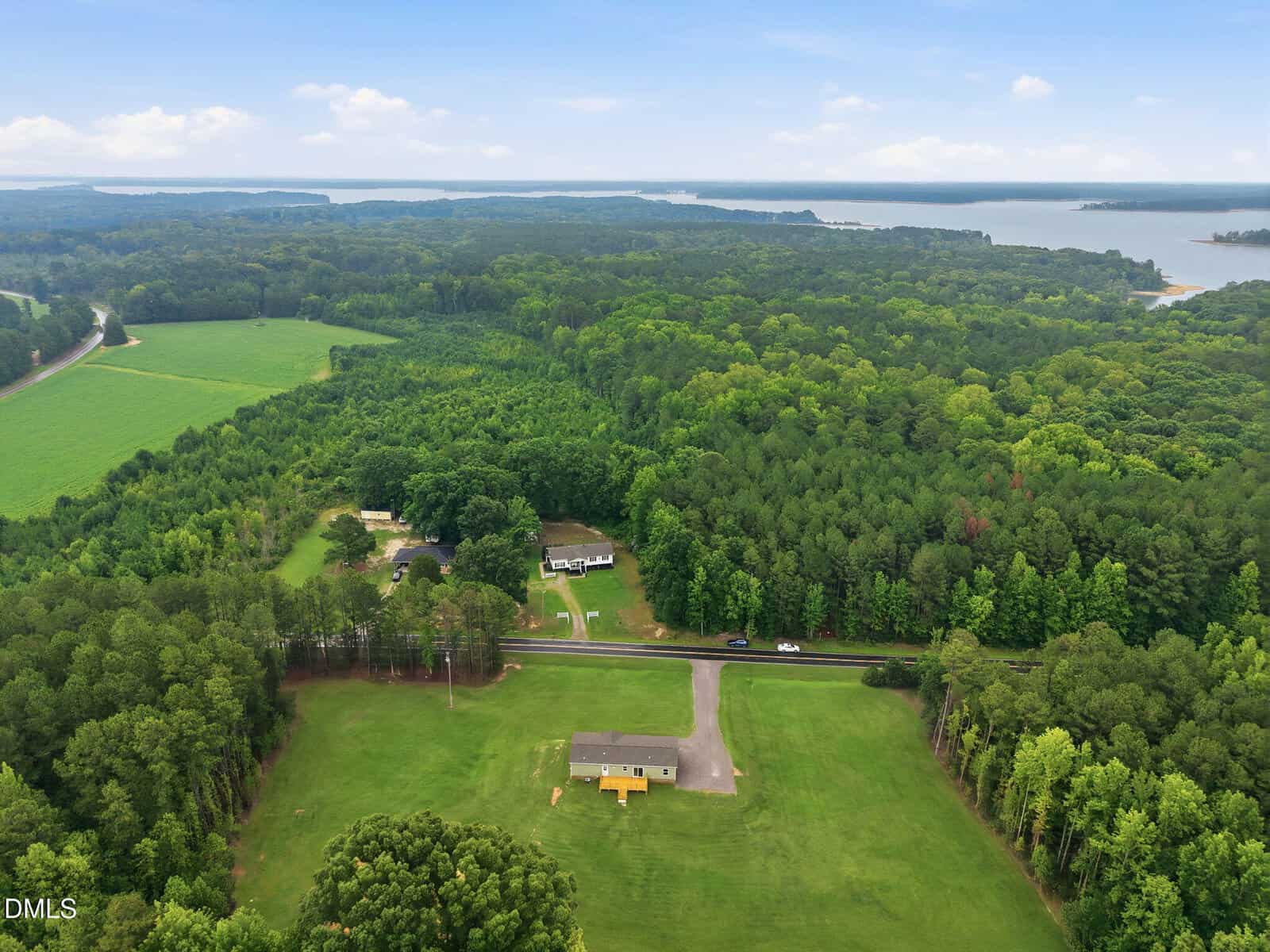Aerial view of a house with a large lawn, surrounded by dense green trees, with a road, other homes, and a lake visible in the background under a partly cloudy sky.