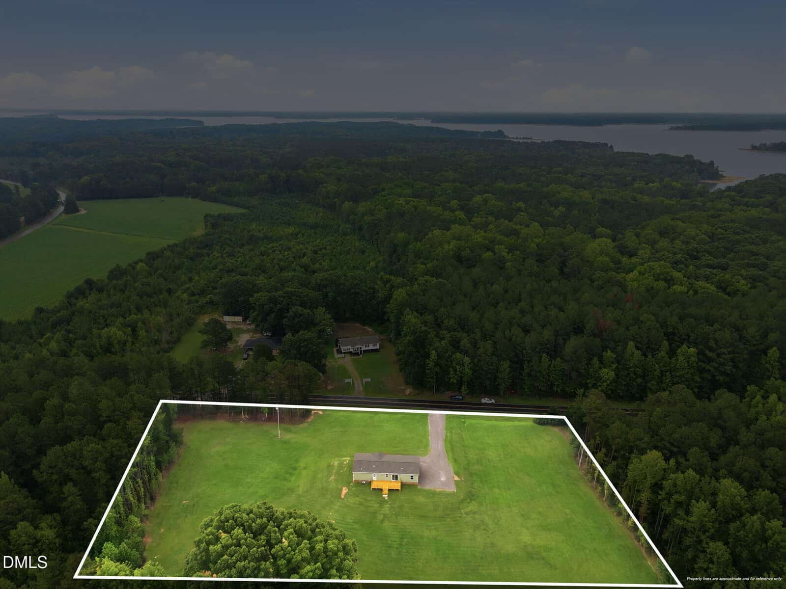 Aerial view of a house on a large green lot outlined in white, surrounded by trees and fields, with a lake visible in the distance under a cloudy sky.