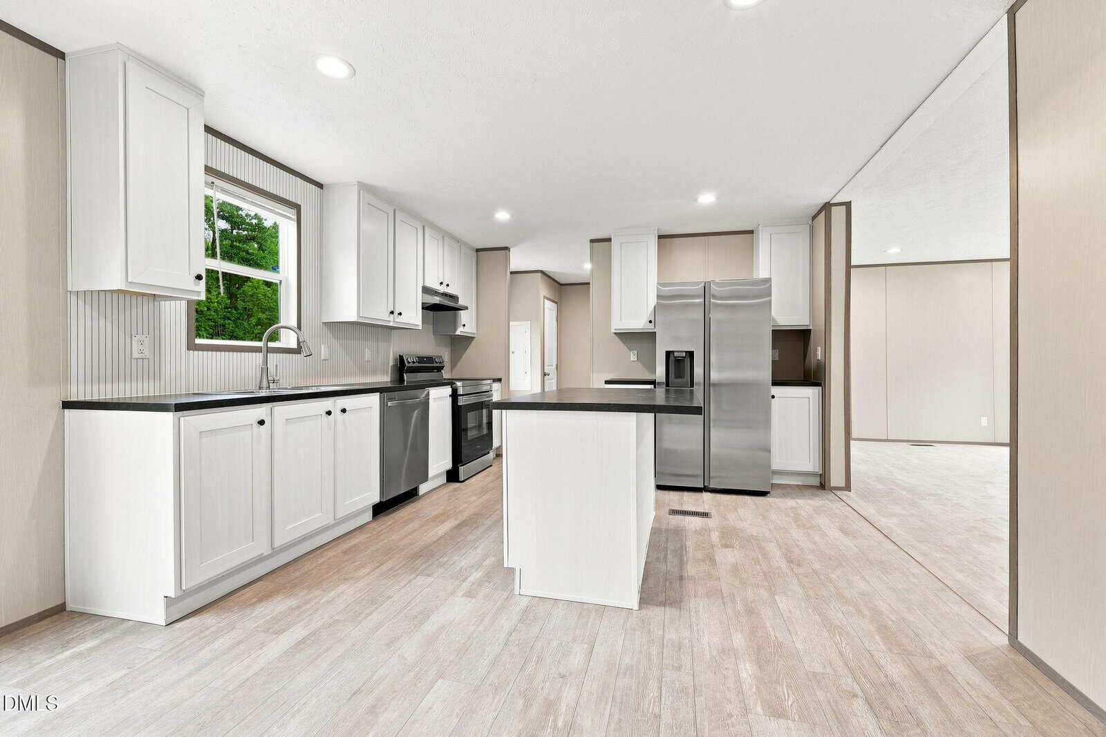 Modern kitchen with white cabinets, stainless steel appliances, small center island, wood-look flooring, and a window above the sink. The space is bright, open, and connects to a larger room.