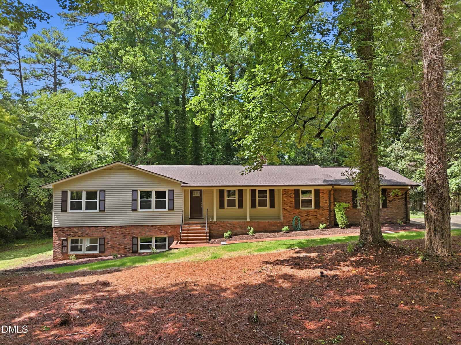 A single-story brick and siding house with a brown roof sits among tall trees. Steps lead to the front door. Multiple windows are visible across the front, and the yard is covered in pine needles and shaded by trees.