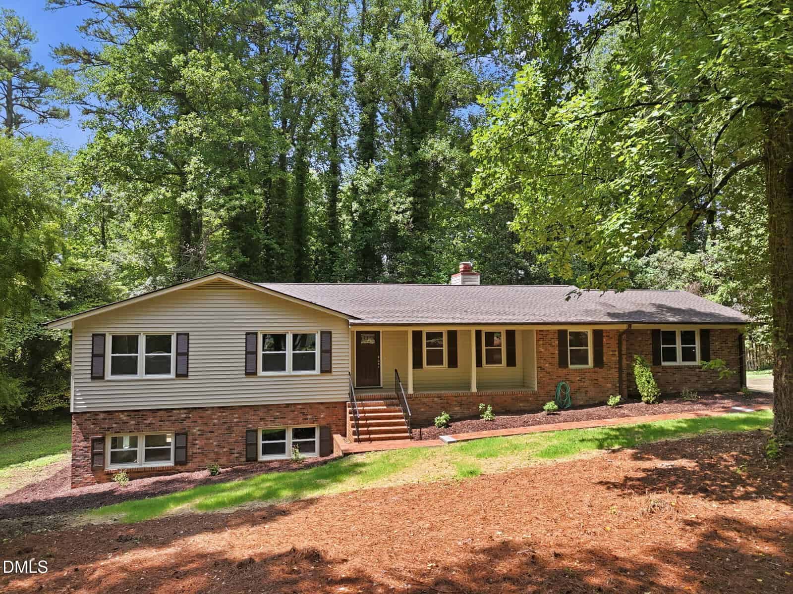 Charming single-story house at 2912 Oxford Road, Henderson, featuring a classic brick and beige siding exterior with dark shutters and a gabled roof. The inviting front porch with steps is surrounded by mature trees, fresh mulch, and a lush grassy lawn under a sunny sky.