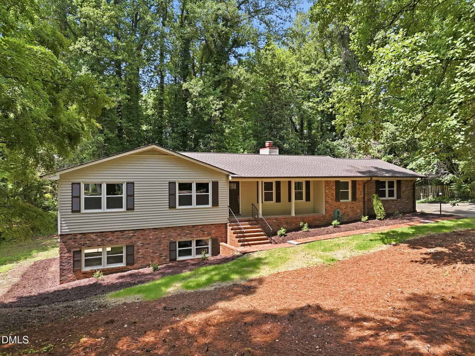 A single-story house with beige siding and brick lower half, brown shutters, and front steps leading to a covered porch, surrounded by tall trees and a mulched yard.