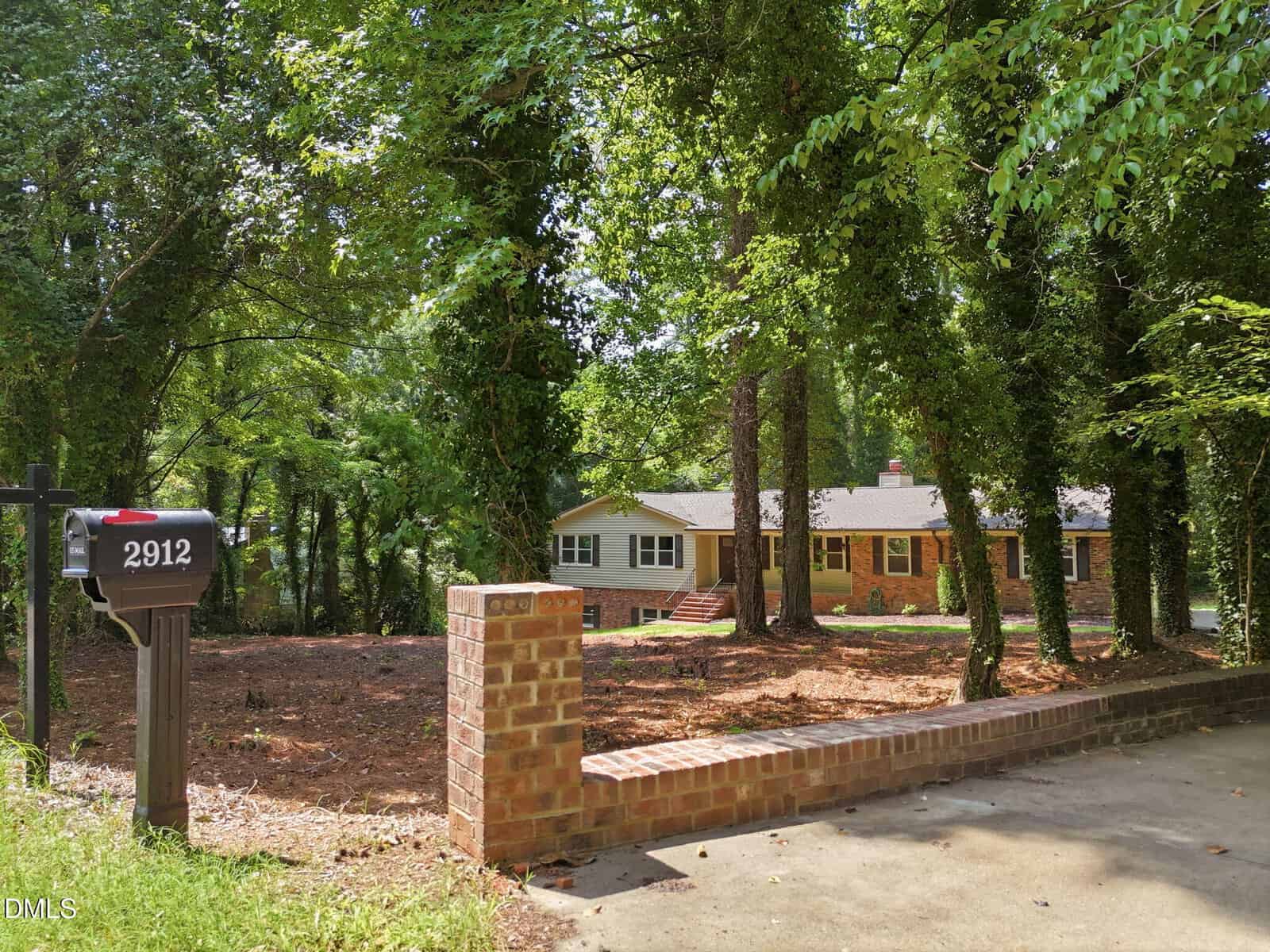 A one-story house with beige siding is partially visible behind tall trees. A brick wall borders the driveway, and a black mailbox with the number 2912 stands at the edge near the street.