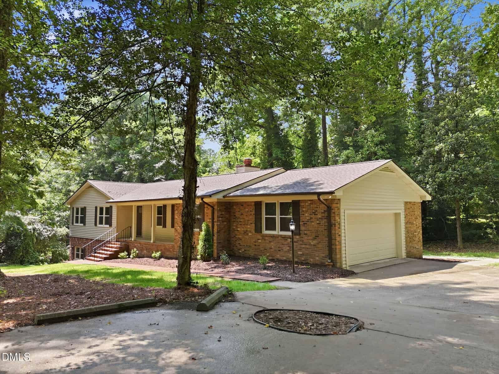 Single-story house with brick and beige siding, attached two-car garage, covered front porch, and a sloped concrete driveway, surrounded by tall trees and greenery.