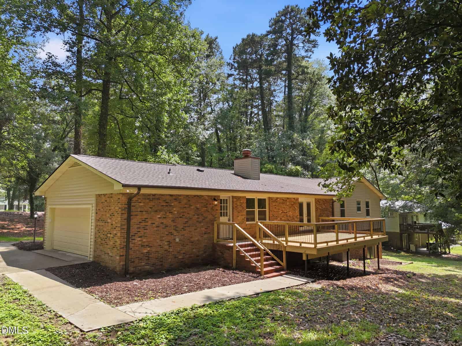 Single-story brick house with attached garage and a wooden deck, surrounded by tall trees and greenery. Concrete driveway leads to the garage. The house is set in a wooded area with ample shade.