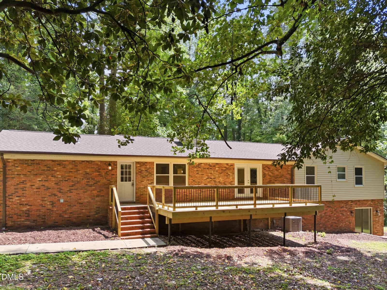 Single-story brick house with a large wooden deck, steps leading to a backyard, and surrounded by green trees and grass on a sunny day.
