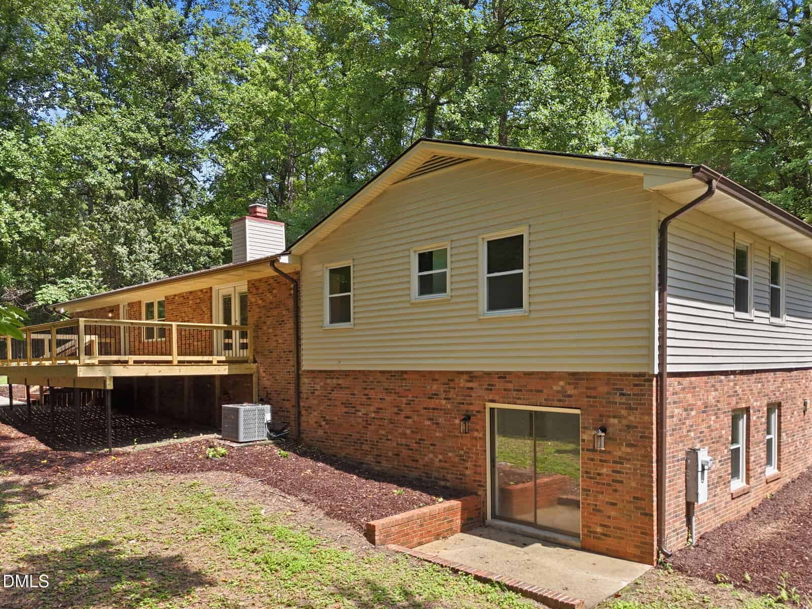 A two-story house with brick and beige siding, multiple windows, a wooden deck, and sliding glass doors, surrounded by trees and greenery.