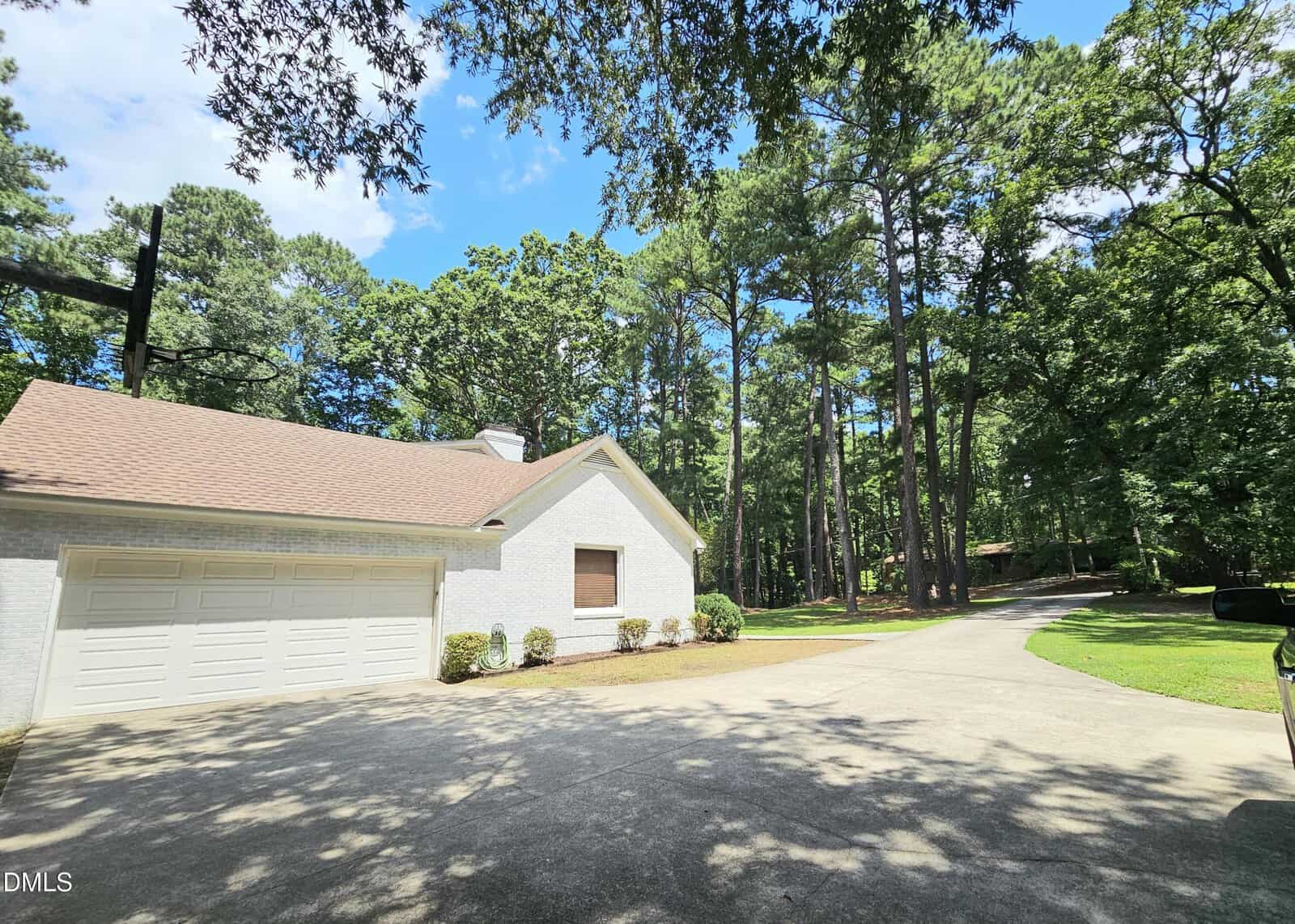 A white house with a two-car garage and brown roof sits beside a curved driveway, surrounded by tall green trees and a well-maintained lawn under a partly cloudy sky.