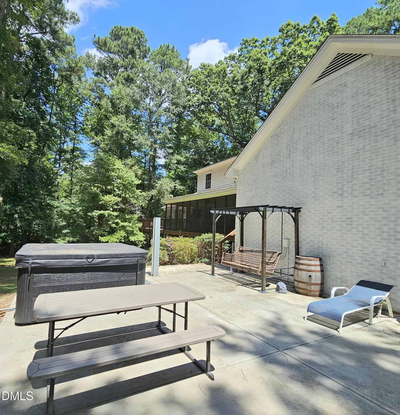 A concrete patio with a picnic table, hot tub, wooden swing under a pergola, barrel, and lounge chair. The patio is next to a light brick house, with trees and greenery in the background under a blue sky.