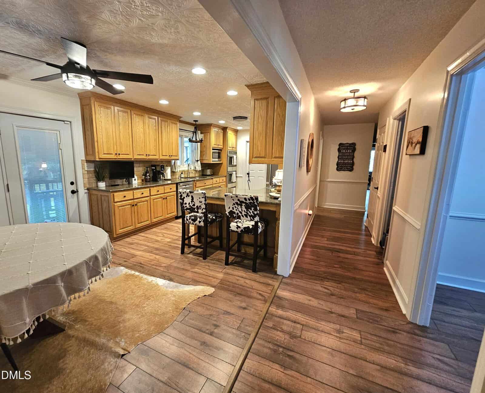Open kitchen with wood cabinets, central island with two patterned chairs, and adjacent dining area. Hallway with wood flooring and ceiling lights leads to several rooms. Bright, inviting, and tidy home interior.