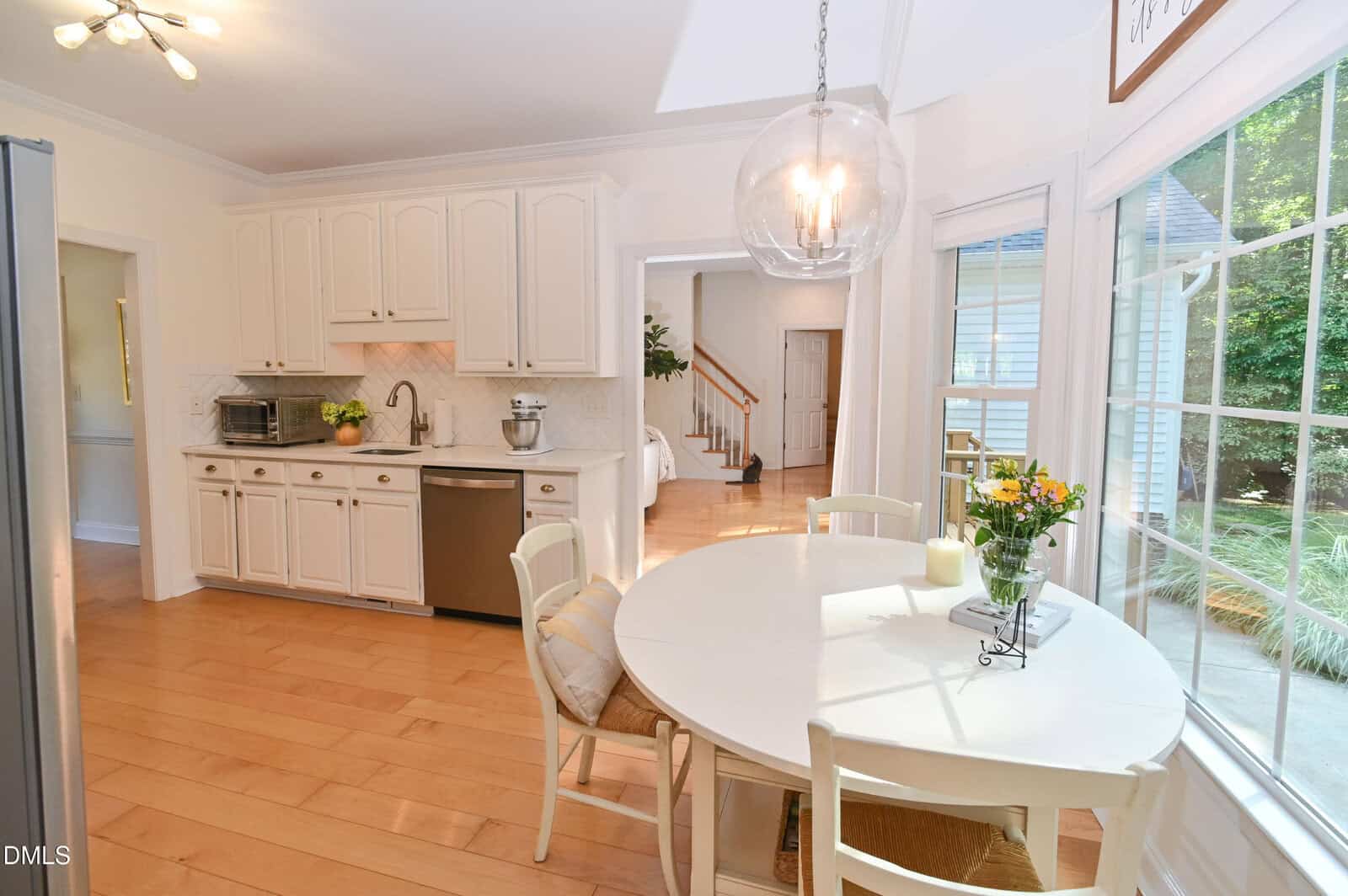 Bright kitchen with white cabinets, light wood flooring, a round dining table near large windows, and a hanging glass light fixture. A vase of flowers sits on the table, and sunlight streams in from outside.