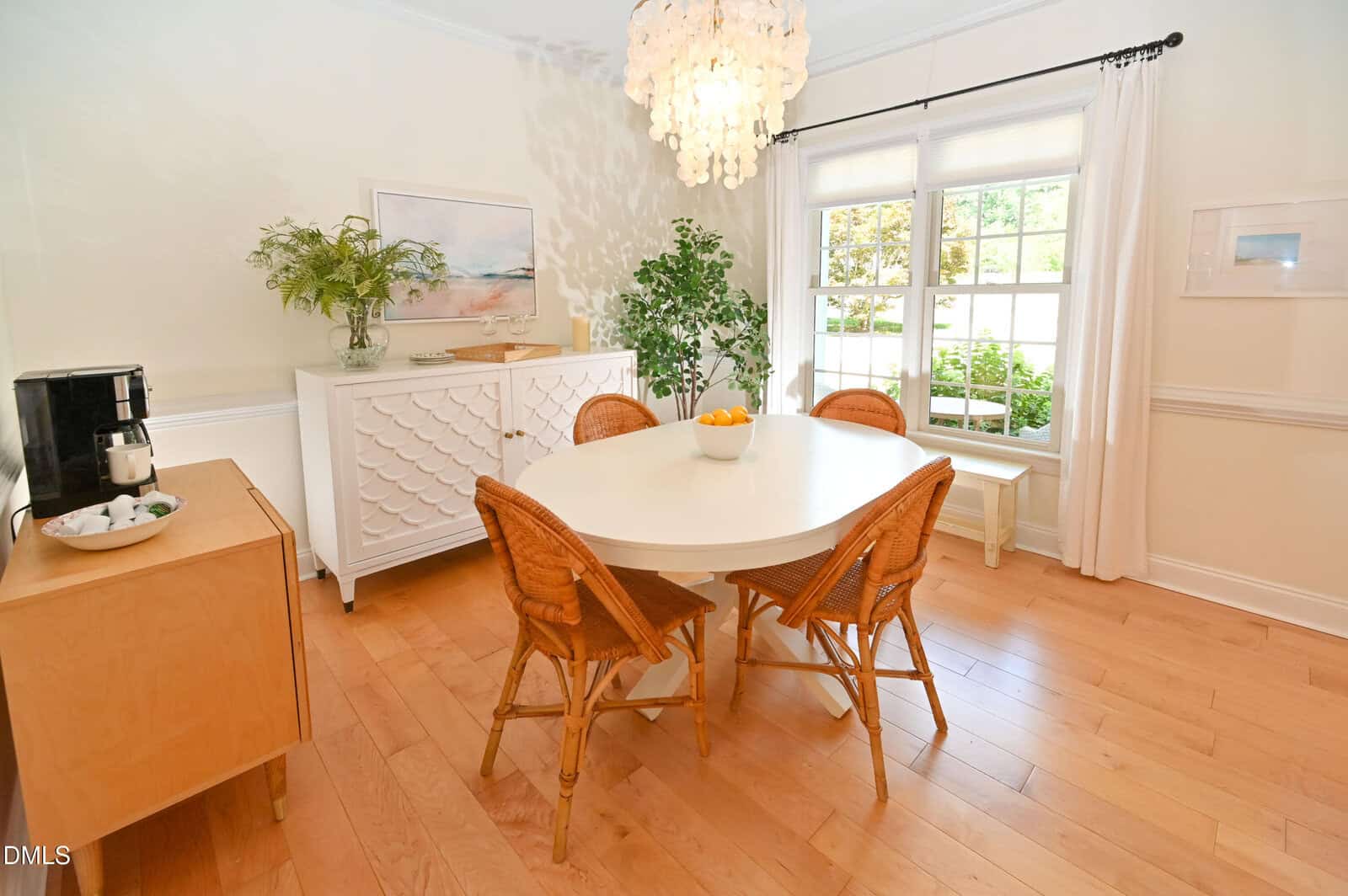 Bright dining area with a round white table, four wicker chairs, a sideboard, potted plants, a coffee maker on a small cabinet, wood floors, and large window with white curtains letting in natural light.