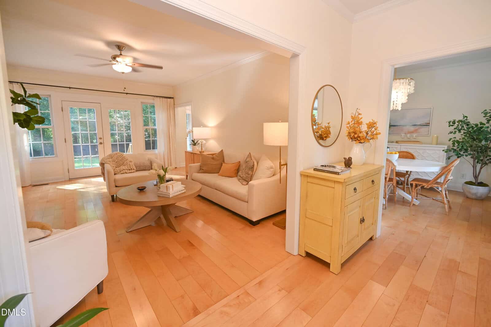 A living room with light wood floors, cream sofas, a round coffee table, and a ceiling fan opens to a dining room with a wooden table and chairs. Large windows let in natural light. A yellow sideboard with a round mirror stands nearby.
