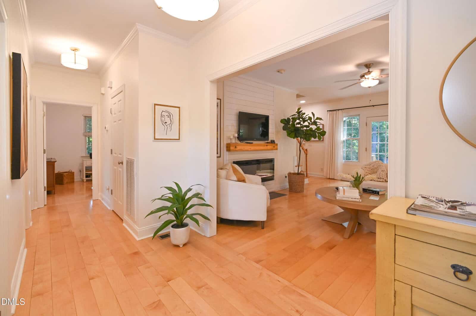 A hallway with light wood floors leads to a living room with a white sofa, wall-mounted TV, round coffee table, potted plant, and large windows with curtains. A yellow console and round mirror are visible on the right.