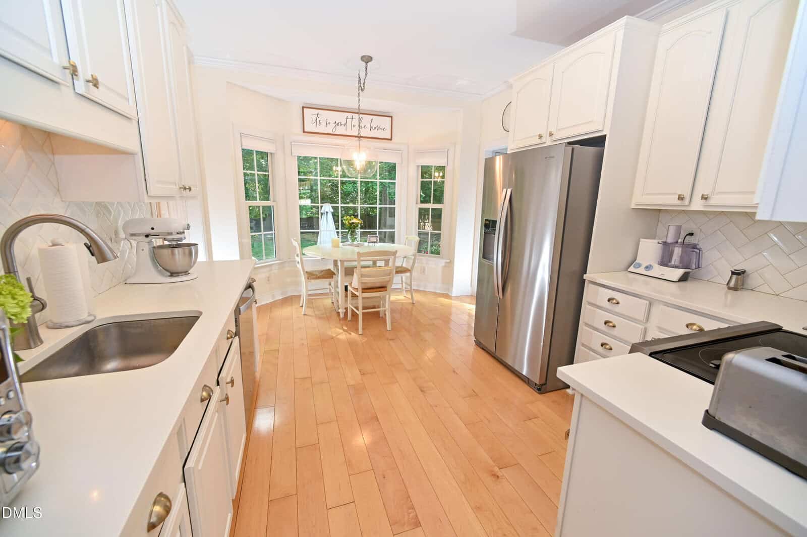 Bright kitchen with white cabinets, stainless steel appliances, wooden floor, and a small dining table by a large bay window with a sign above. Mixing bowls and appliances are on the counters.