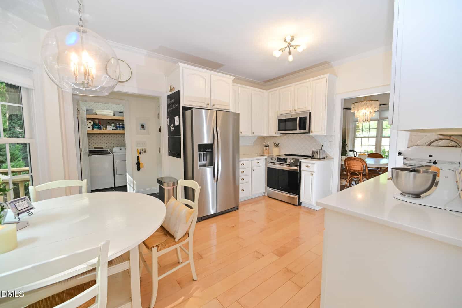Bright kitchen with white cabinets, stainless steel appliances, a round dining table with four chairs, and hardwood floors. An adjacent laundry room and dining area are visible through doorways.
