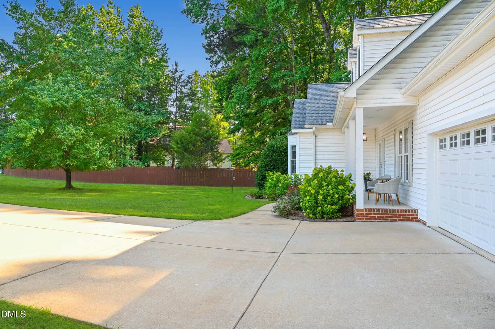 A white house with an attached garage, a small front porch with chairs, a wide driveway, and a lawn bordered by trees and a wooden fence under a clear blue sky.