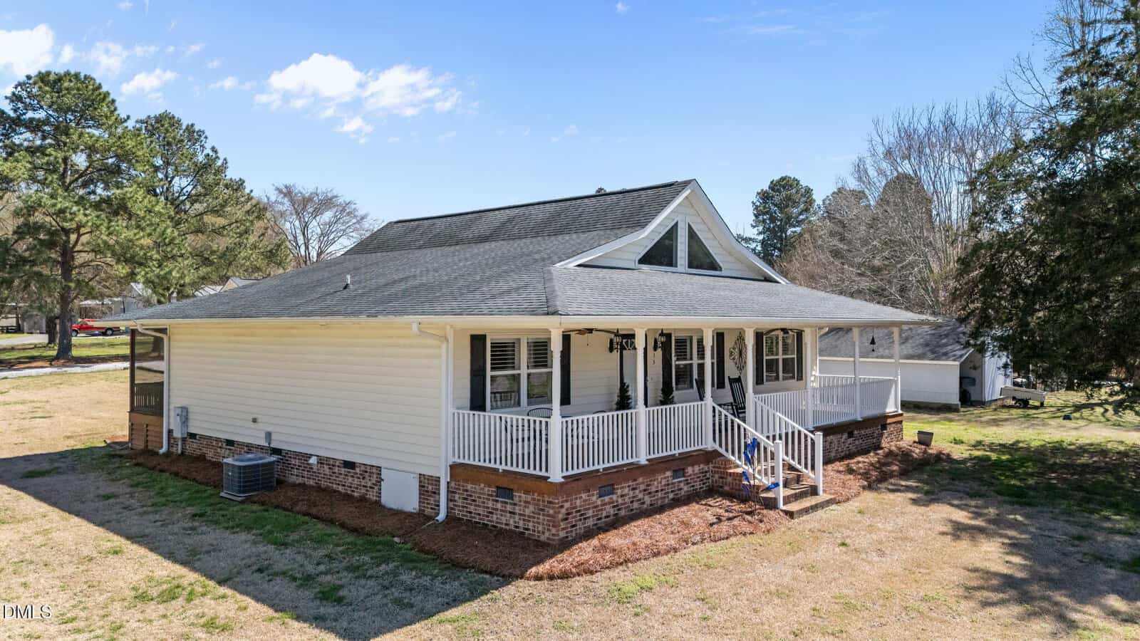 Single-story house with white siding, dark roof, brick foundation, and covered front porch with railings. The yard is open with grass patches, trees in the background, and a blue sky with some clouds above.