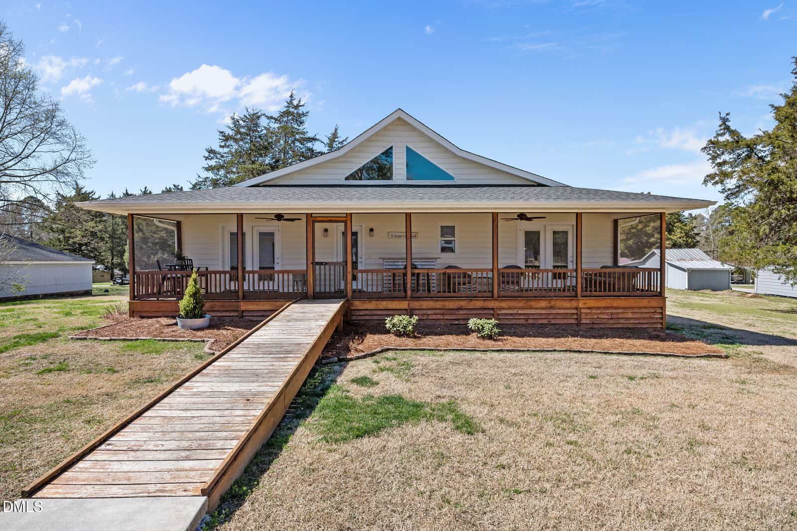 A single-story white house with a triangular window, wide front porch, wheelchair-accessible wooden ramp, screened sections, and a neatly trimmed lawn under a blue sky. Trees and a small shed are visible in the background.