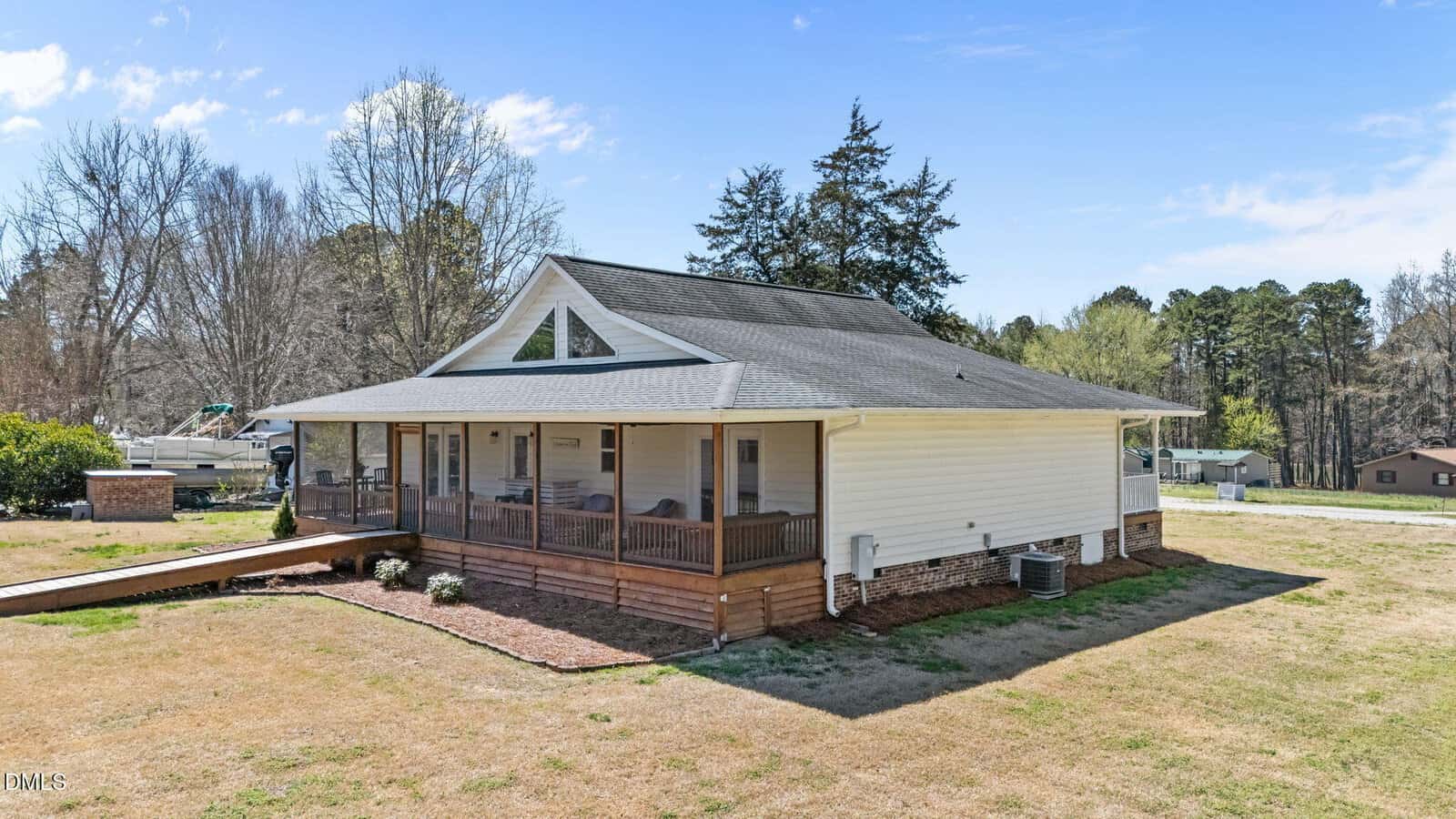 Single-story house with white siding and a gray roof, featuring a large screened-in porch with wooden trim and a ramp. The house is surrounded by a grassy yard with trees and other homes visible in the background.