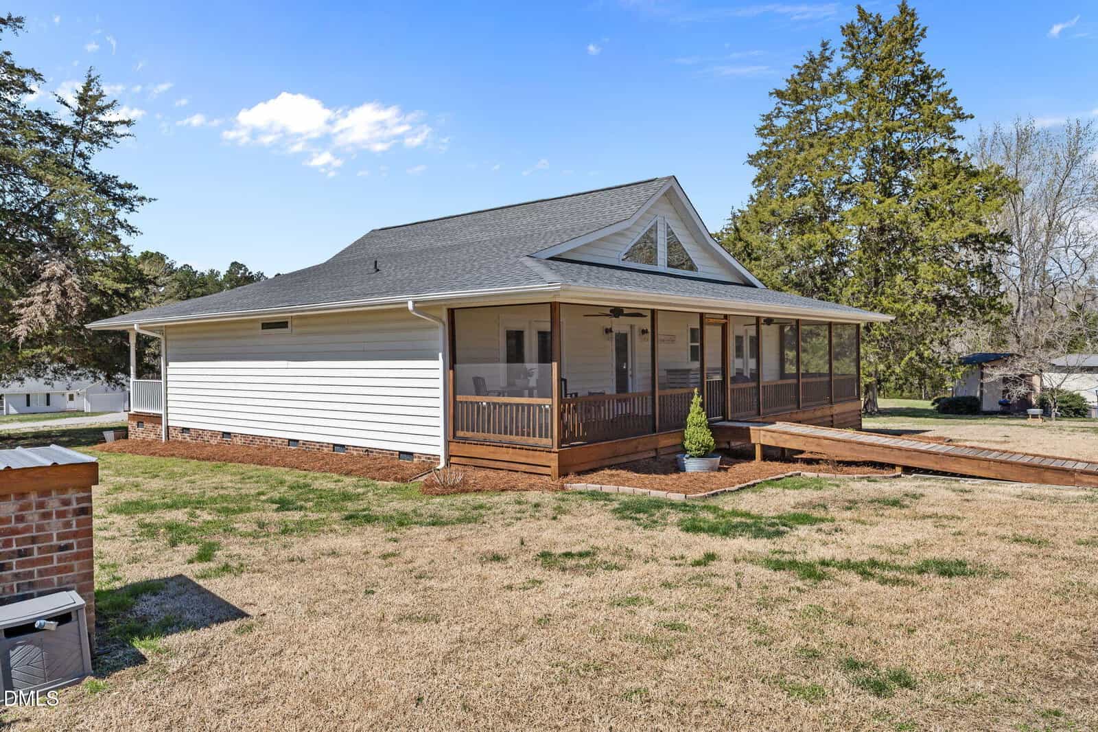 A single-story house with white siding, a dark roof, and a large wraparound screened porch. The yard is grassy with some trees in the background, and there is a wooden ramp leading to the porch.