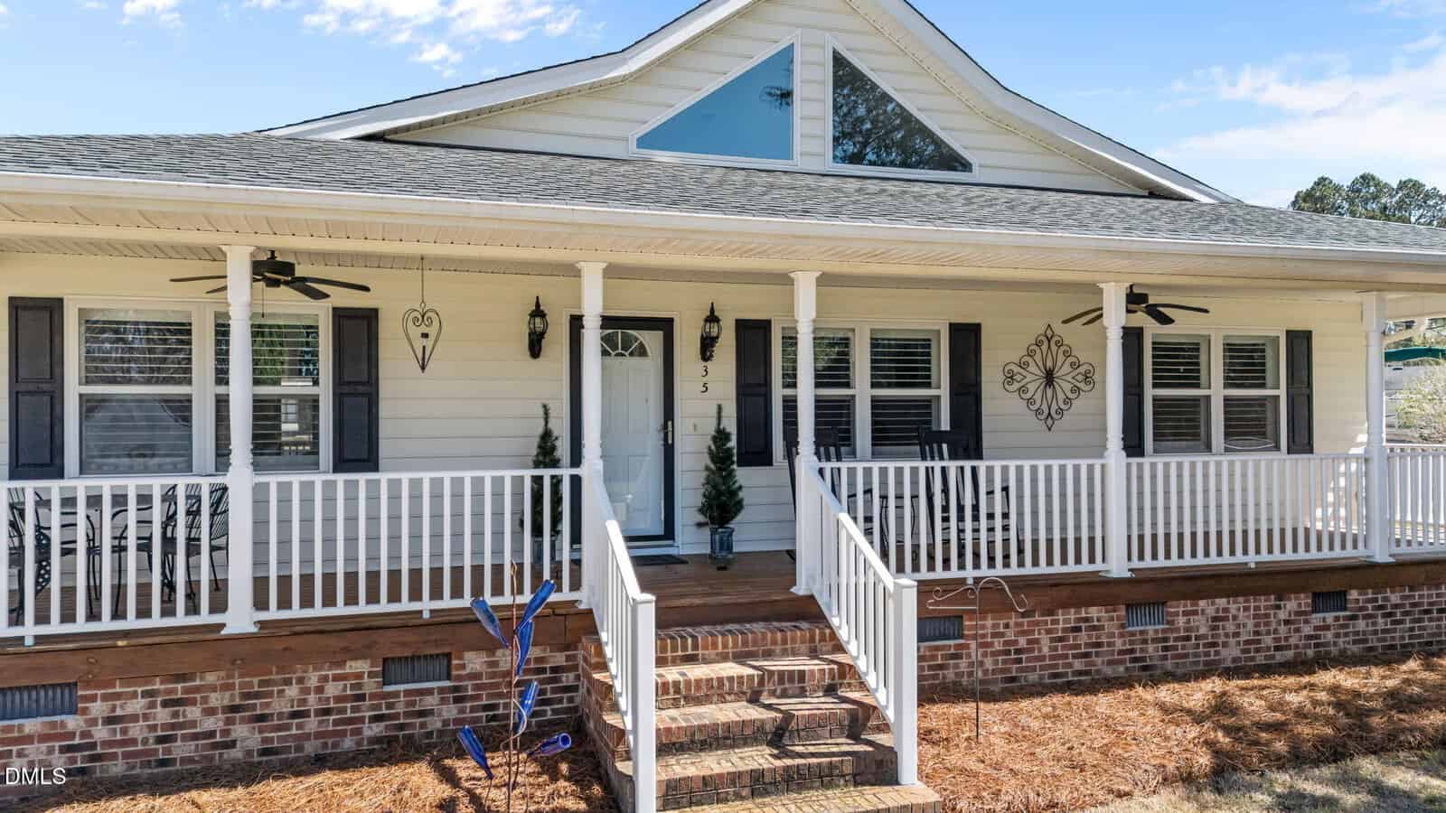 A white house with a large front porch, brick steps, white railings, two ceiling fans, and decorative wall art. There are two small trees by the door and a blue wind spinner in the yard.