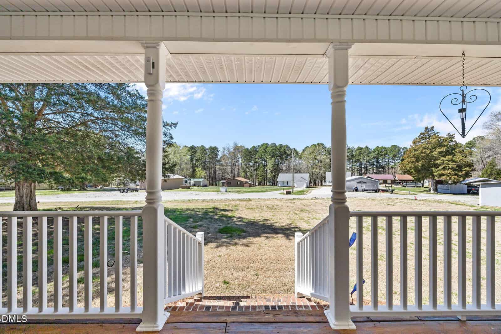 View from a porch with white railings and steps leading to a grassy yard, overlooking a suburban street with houses, trees, and clear blue sky in the background.