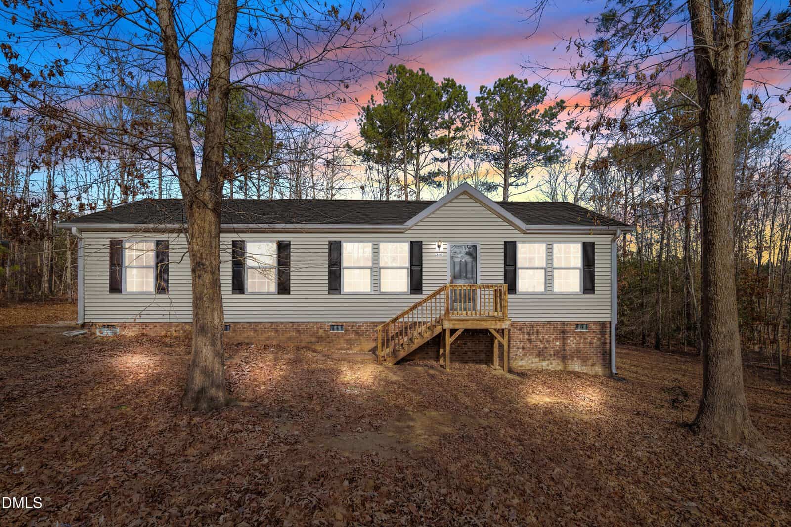 Charming single-story house at 280 Rough Lane, Louisburg, with beige siding, black shutters, and a small wooden front porch. The yard is covered in fallen leaves, while trees surround the property. The sky above glows with colorful sunset hues.