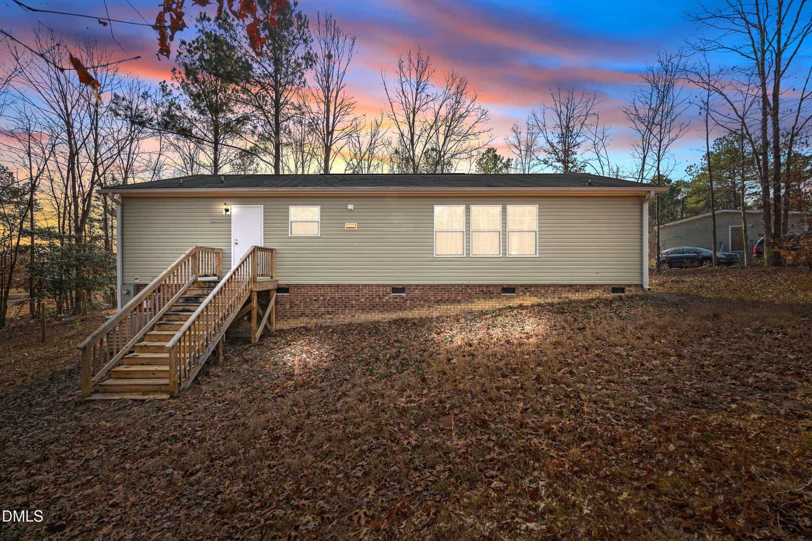 Single-story beige house with a brick foundation, wooden front steps, and white door, situated on a sloped, leaf-covered yard. Bare trees surround the property, and the sky shows sunset colors.