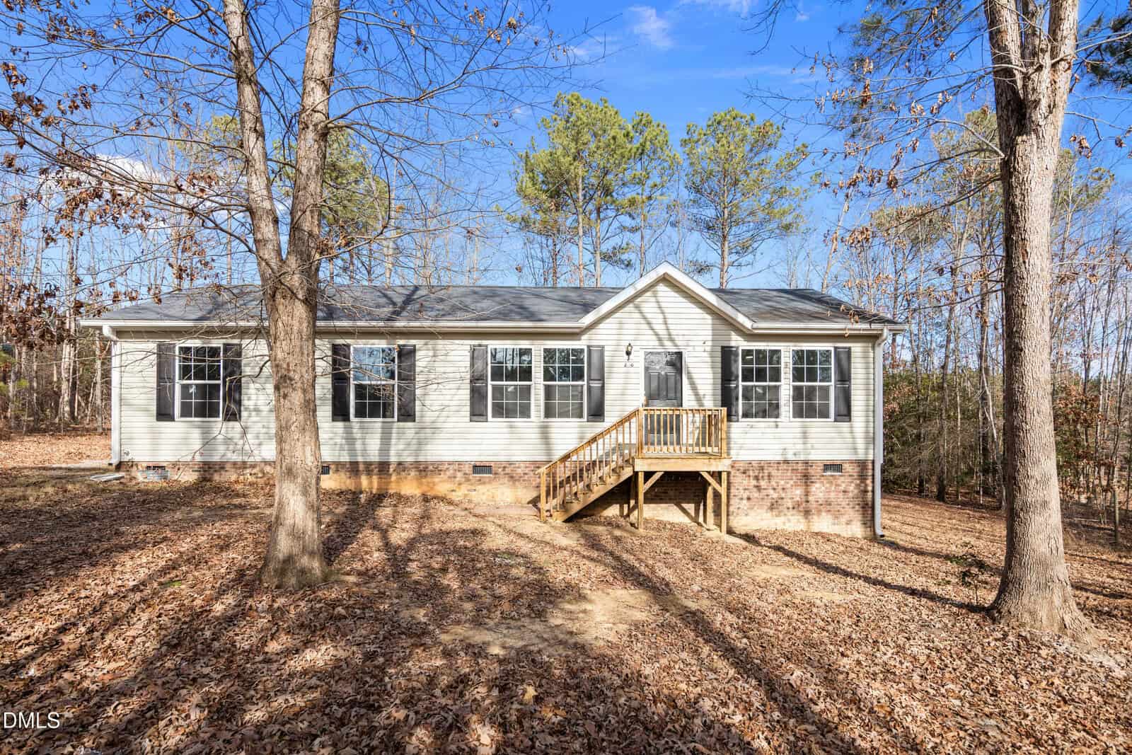 Single-story house with beige siding, black shutters, and a small front porch with wooden steps. Surrounded by leaf-covered ground and tall trees, under a clear blue sky.