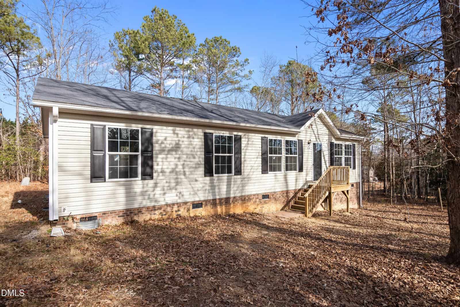 Single-story beige house with black shutters, multiple windows, a small wooden front porch, and steps. The house is surrounded by trees and fallen leaves, with a clear blue sky overhead.